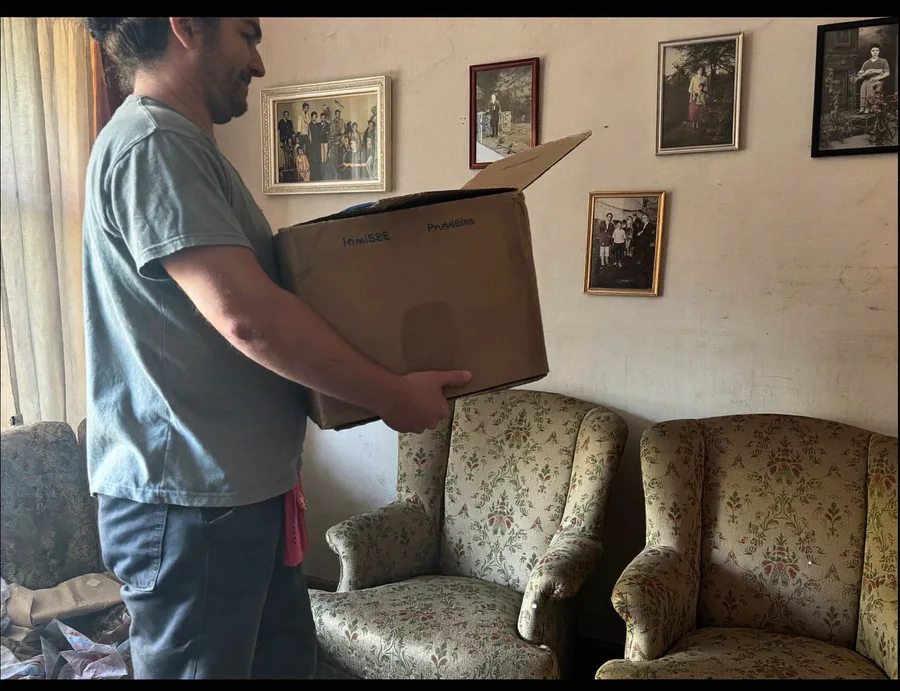 Worker carefully carrying a box past armchairs and family photos during an estate cleanout